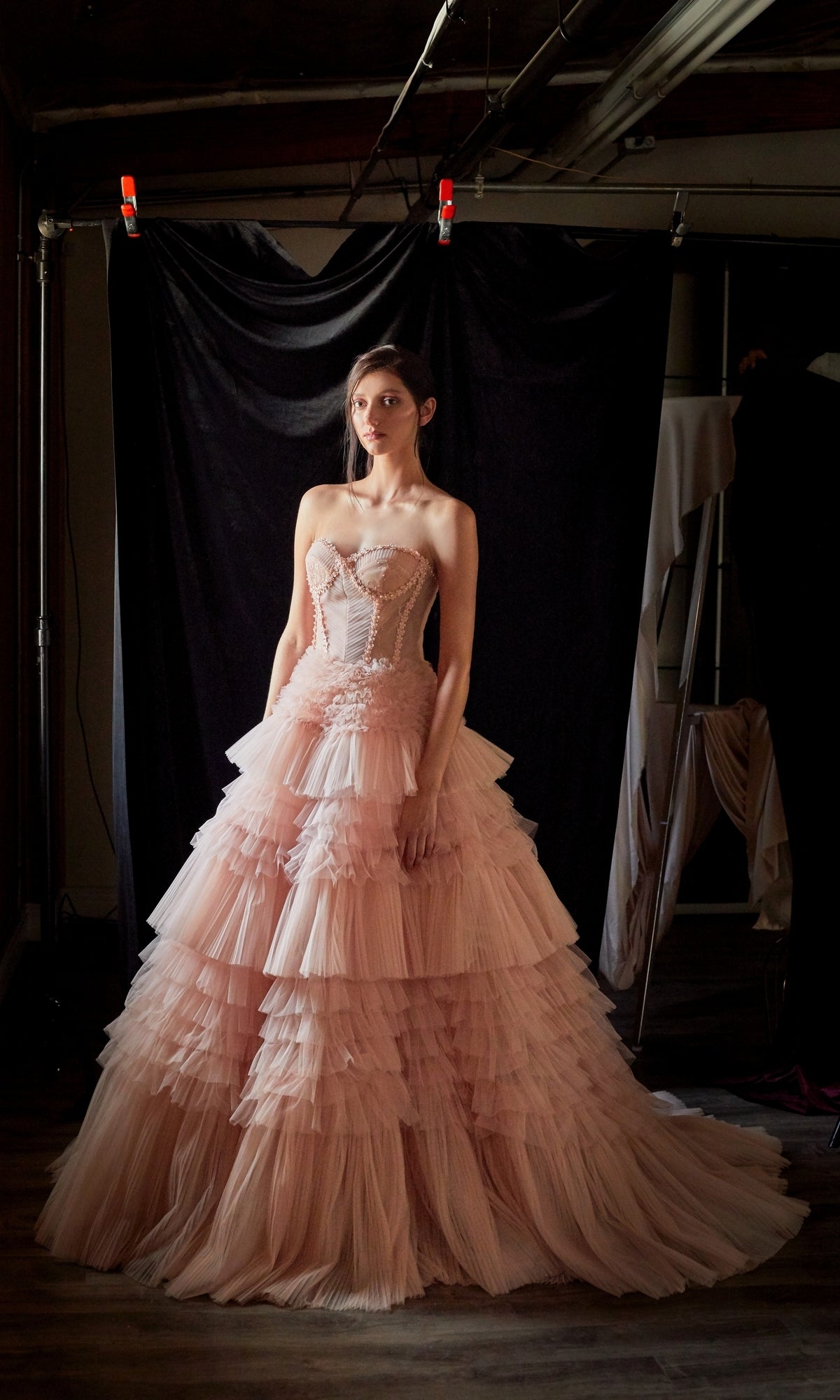 A woman stands before a black backdrop in a dimly lit room, wearing the Strapless Tiered Long Prom Ball Gown A1017, featuring layers of pink ruffles.